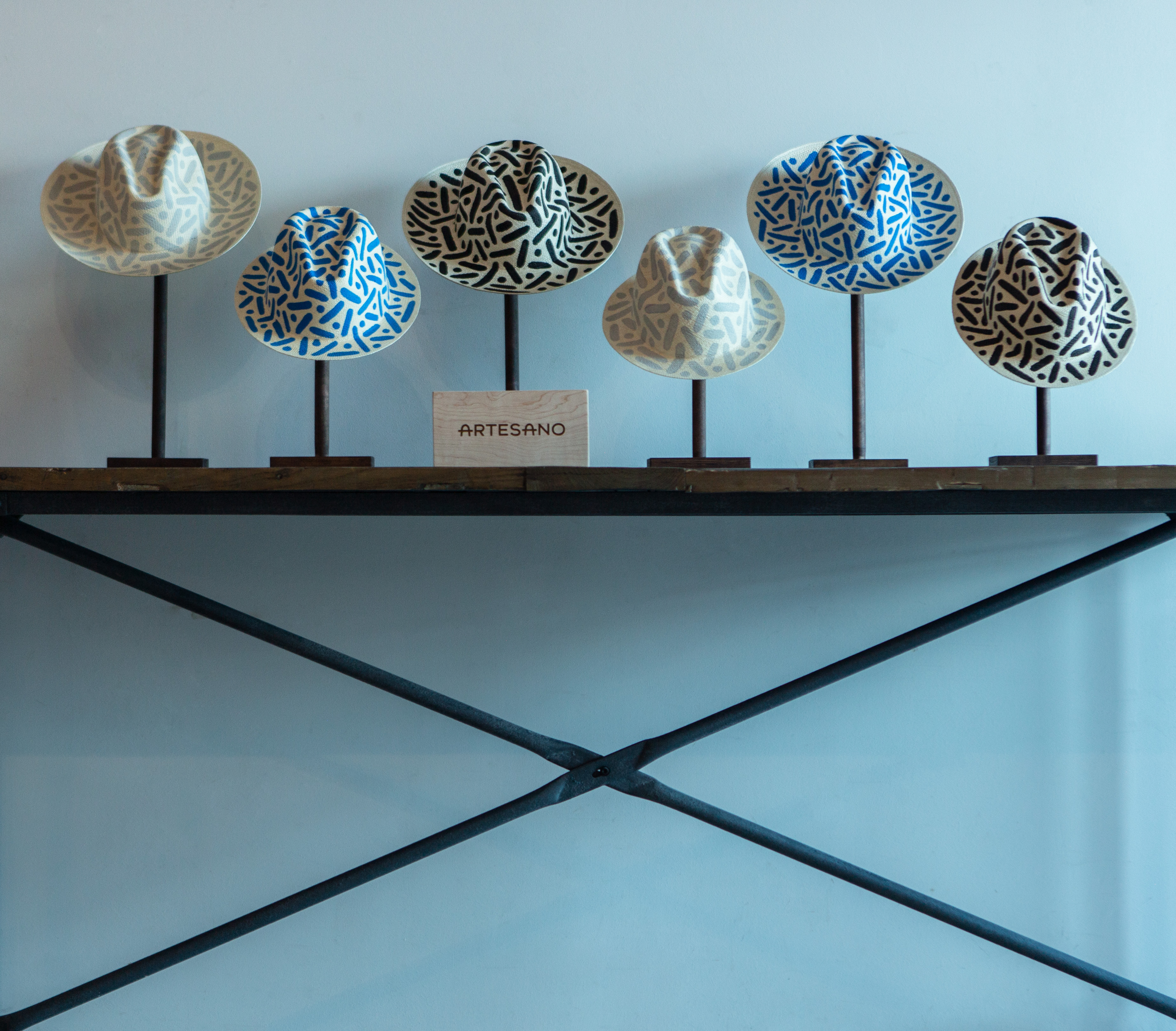 Collection of hand-painted Panama hats from the Artesano × P!PE collaboration, displayed on stands above a wooden table. Each hat features Pipe Yanguas's distinctive dots-and-lines patterns in black, gray, or blue.