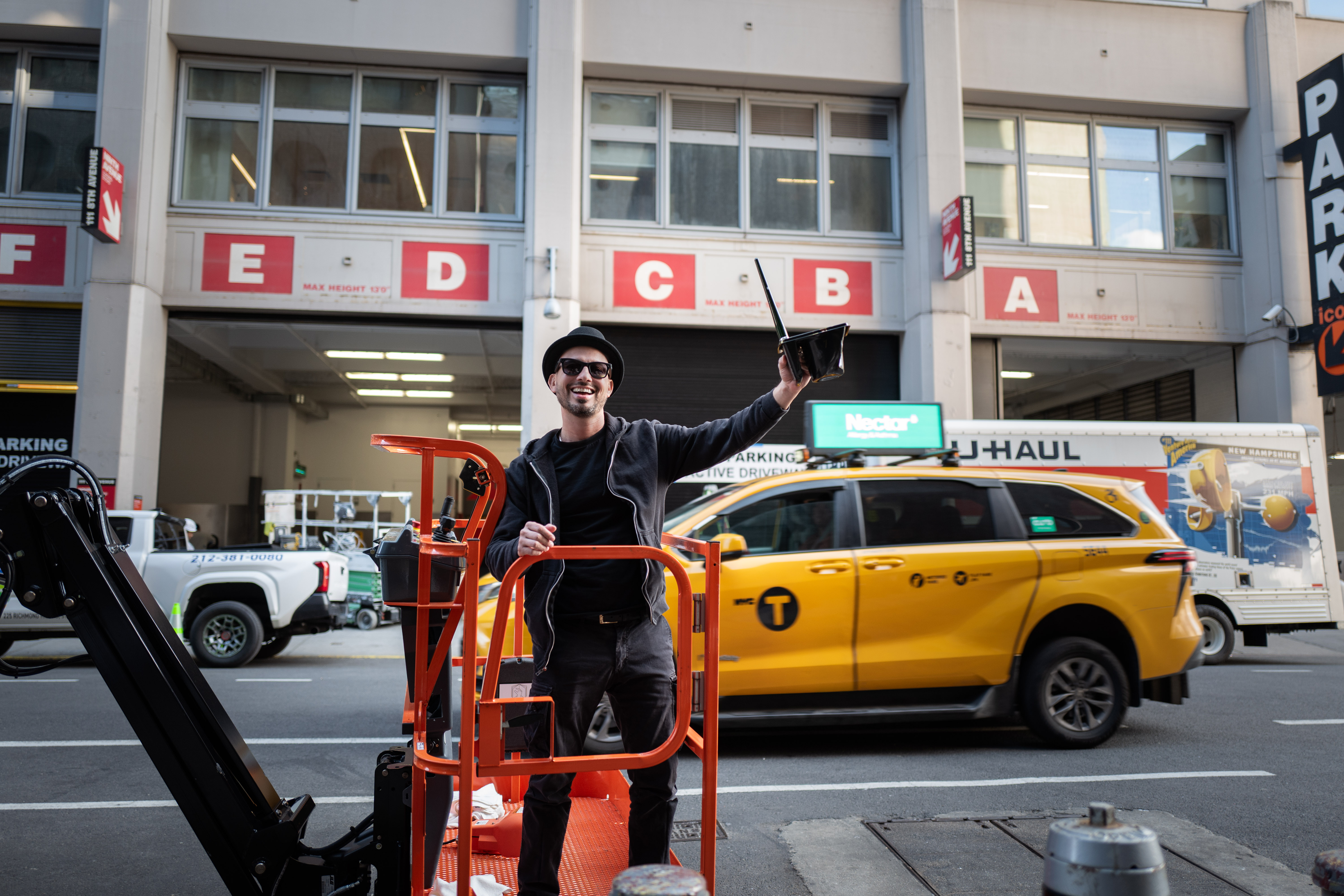 The Golden Weave: Artist stands in an orange lift basket beside a street with a passing yellow taxi.