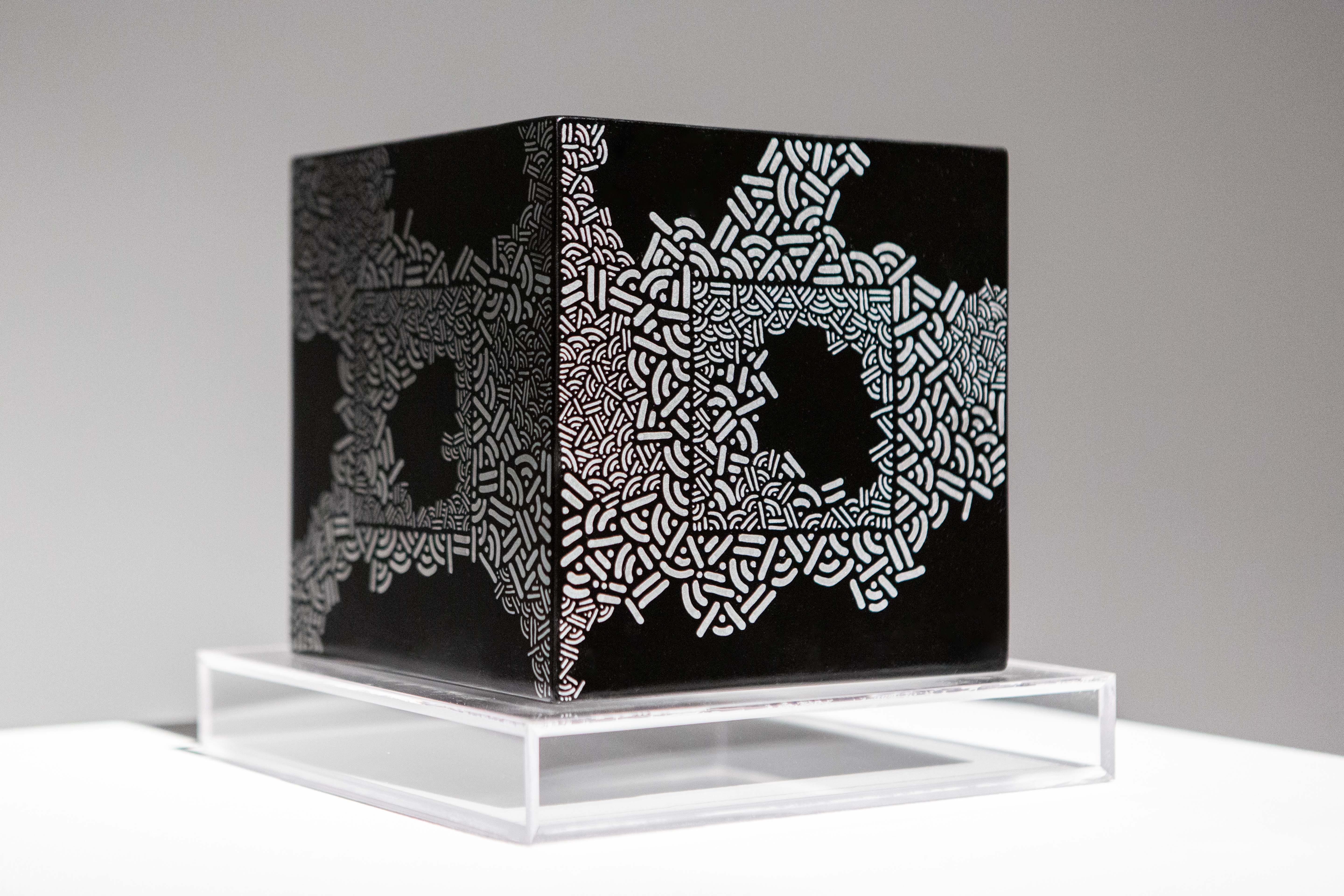 Black cube decorated with pale geometric clusters rests on a clear display stand.