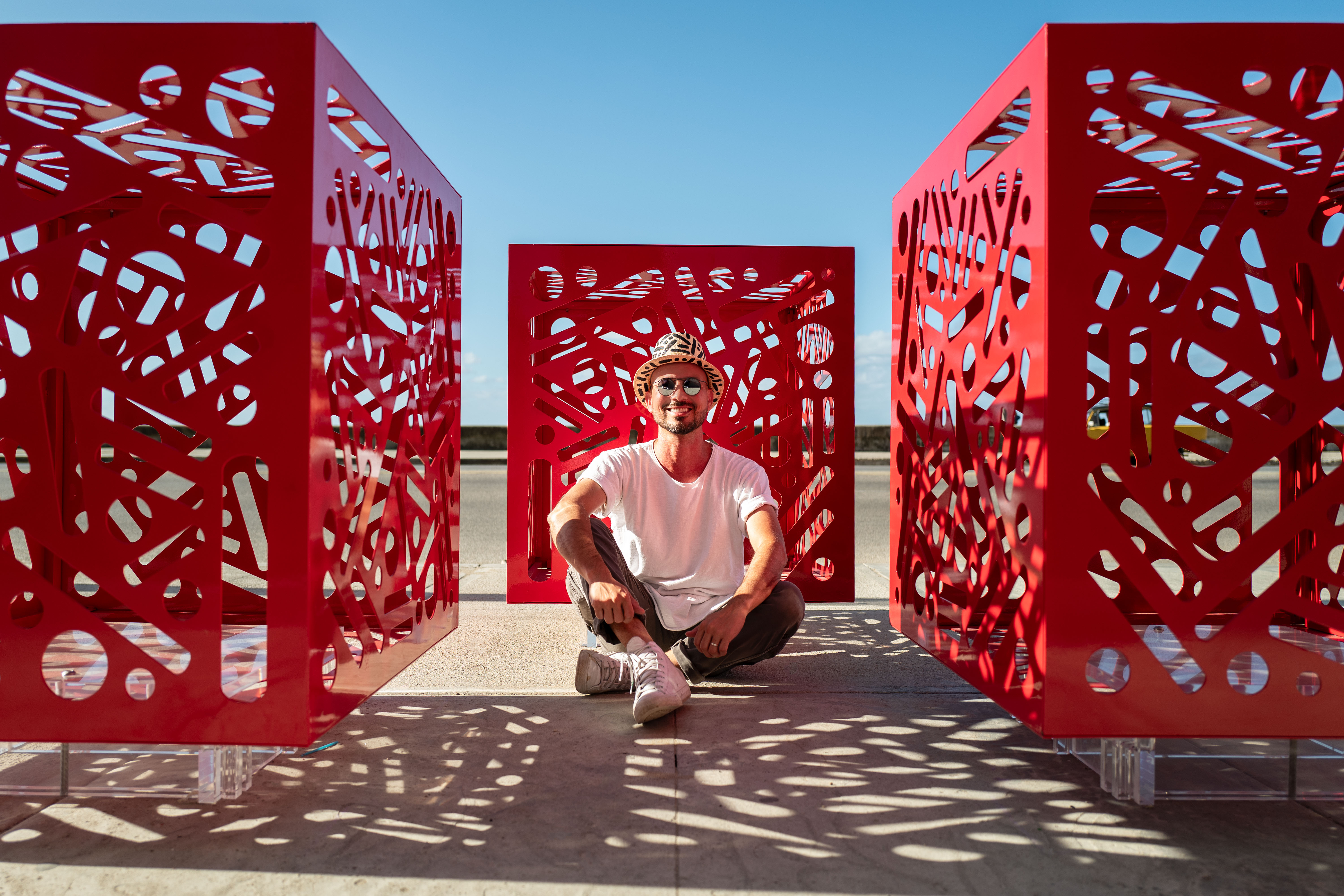 Hemoglobin: Person sits between three red cutout cube sculptures on sunlit pavement.