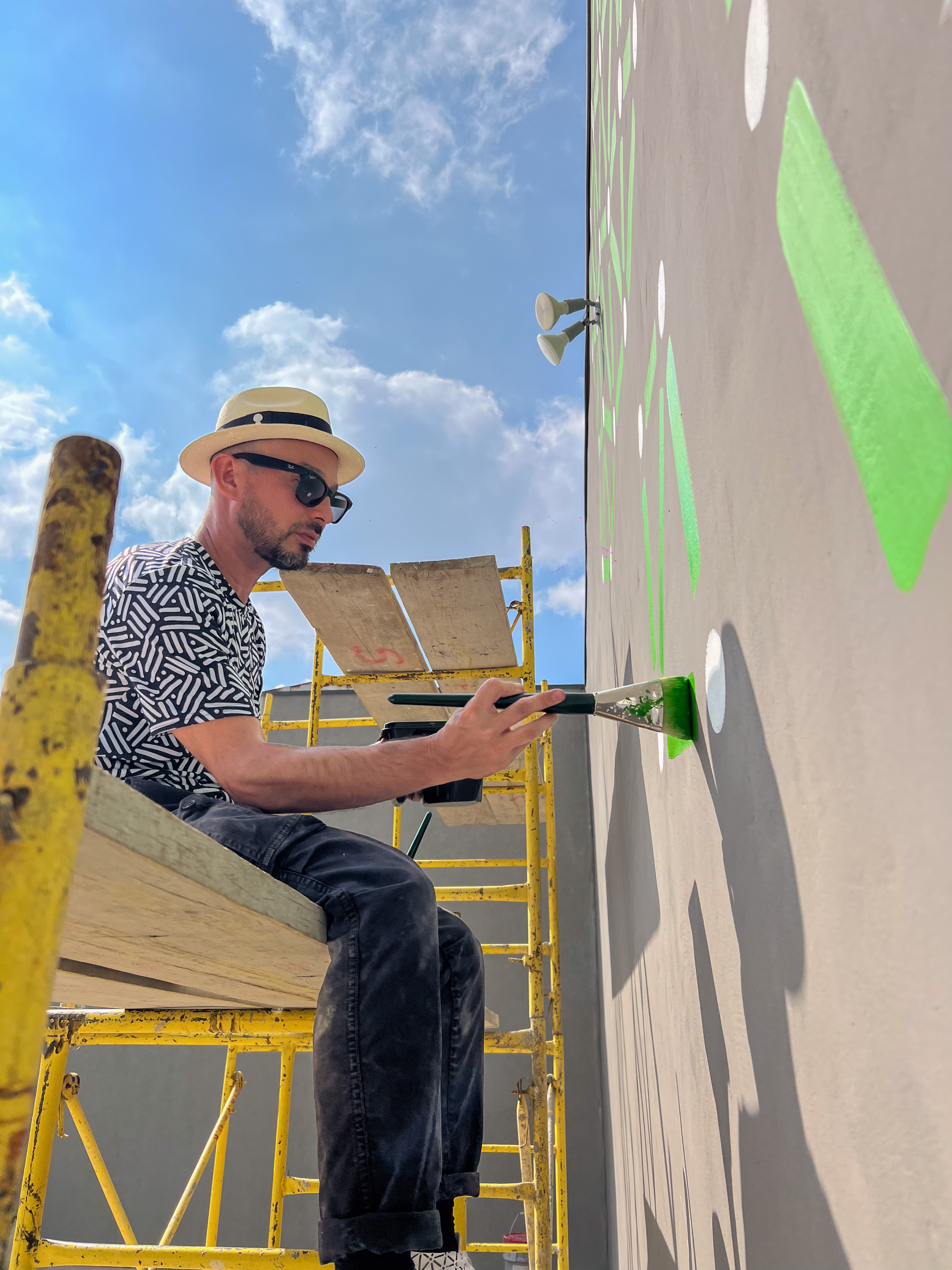 Roots: Artist sits on yellow scaffolding and paints green marks on a gray courtyard wall.