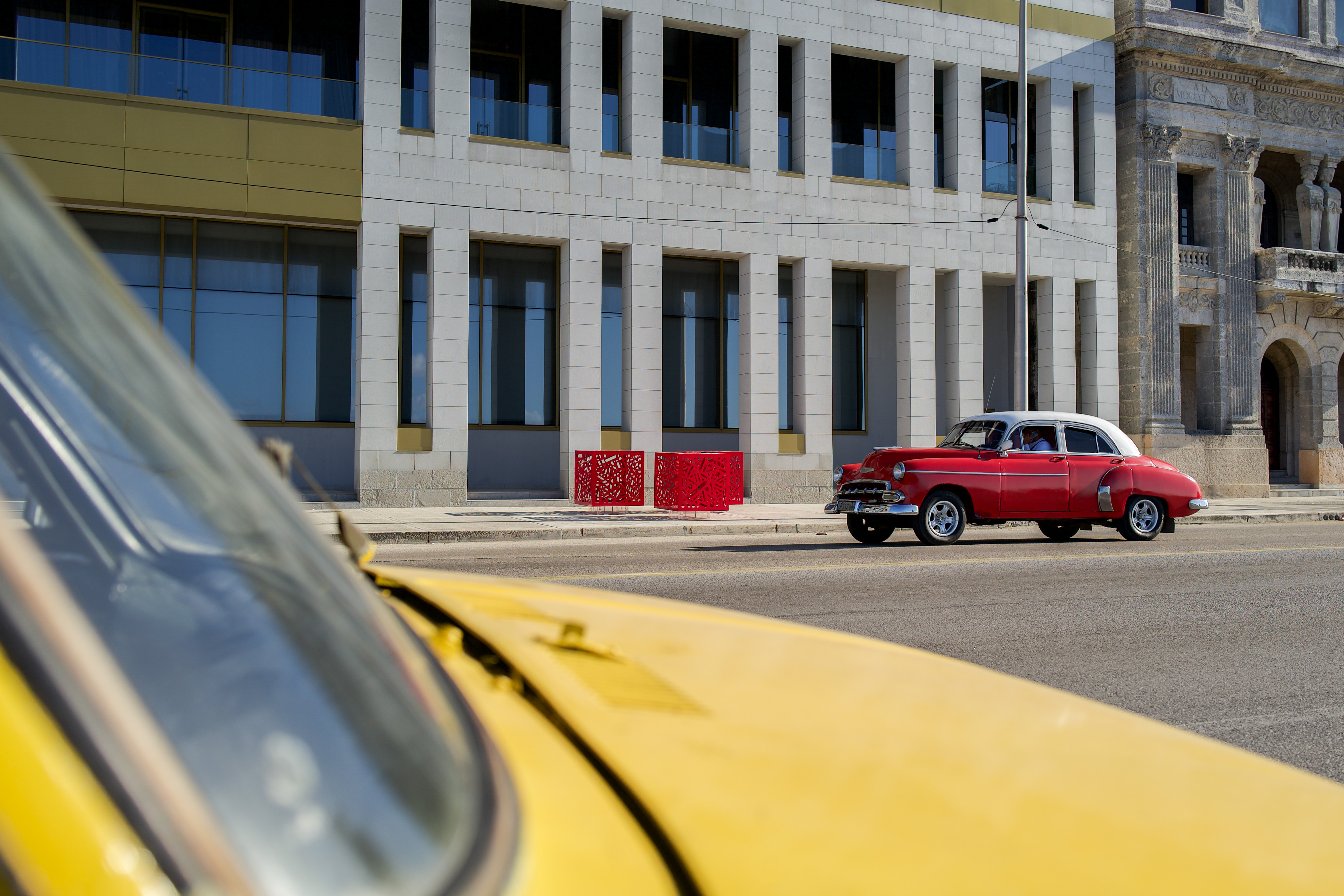 Hemoglobin - Installation of 3: Three red cutout cube sculptures on a city sidewalk beside a road and a large building.