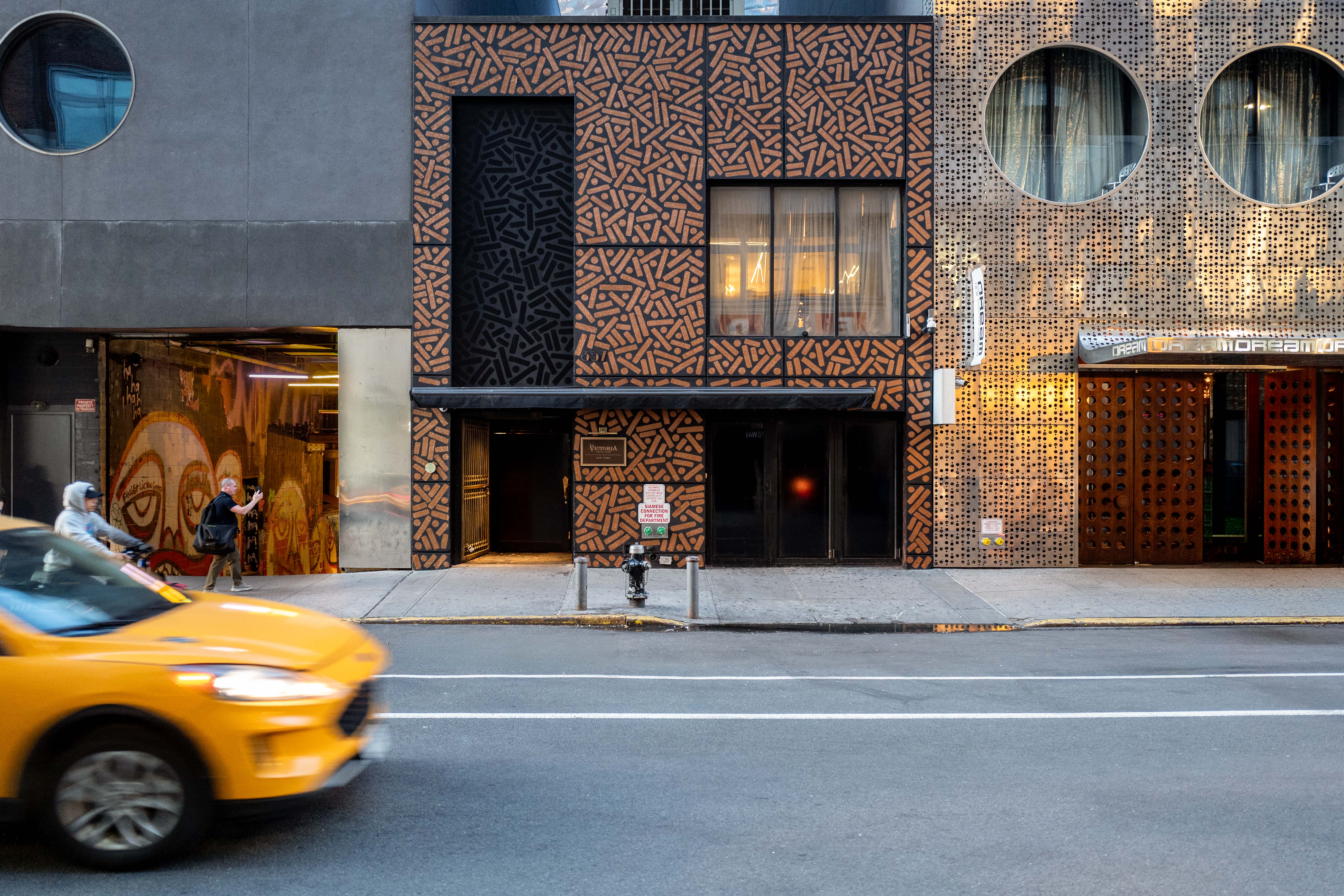 Street view of a dark storefront and a golden patterned facade, with a yellow taxi passing in front.
