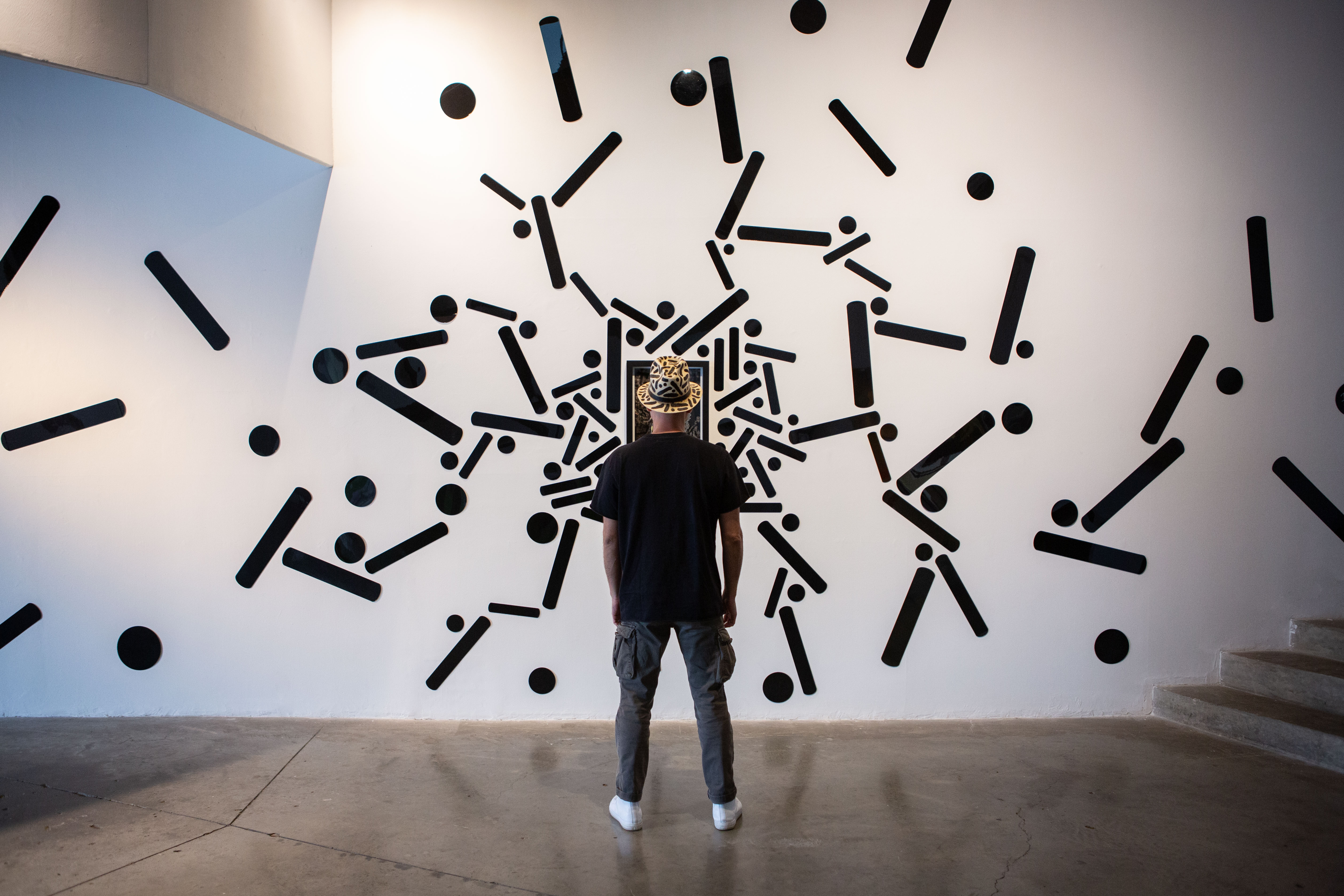 Pipe Yanguas, seen from behind in a patterned hat, stands before a white wall covered in large black dots and lines.