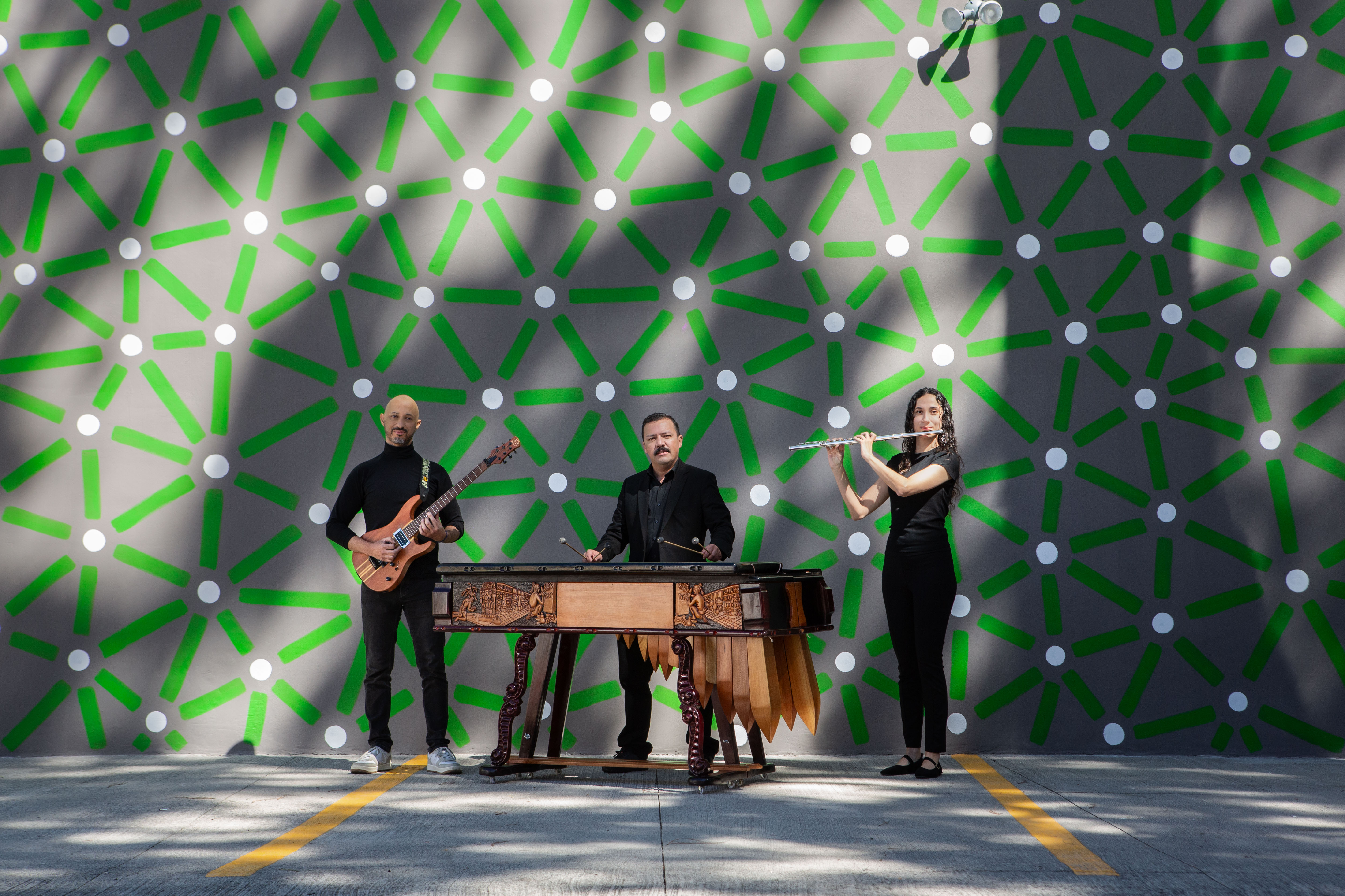 Three musicians perform in front of a wall patterned with green connecting lines and pale dots.