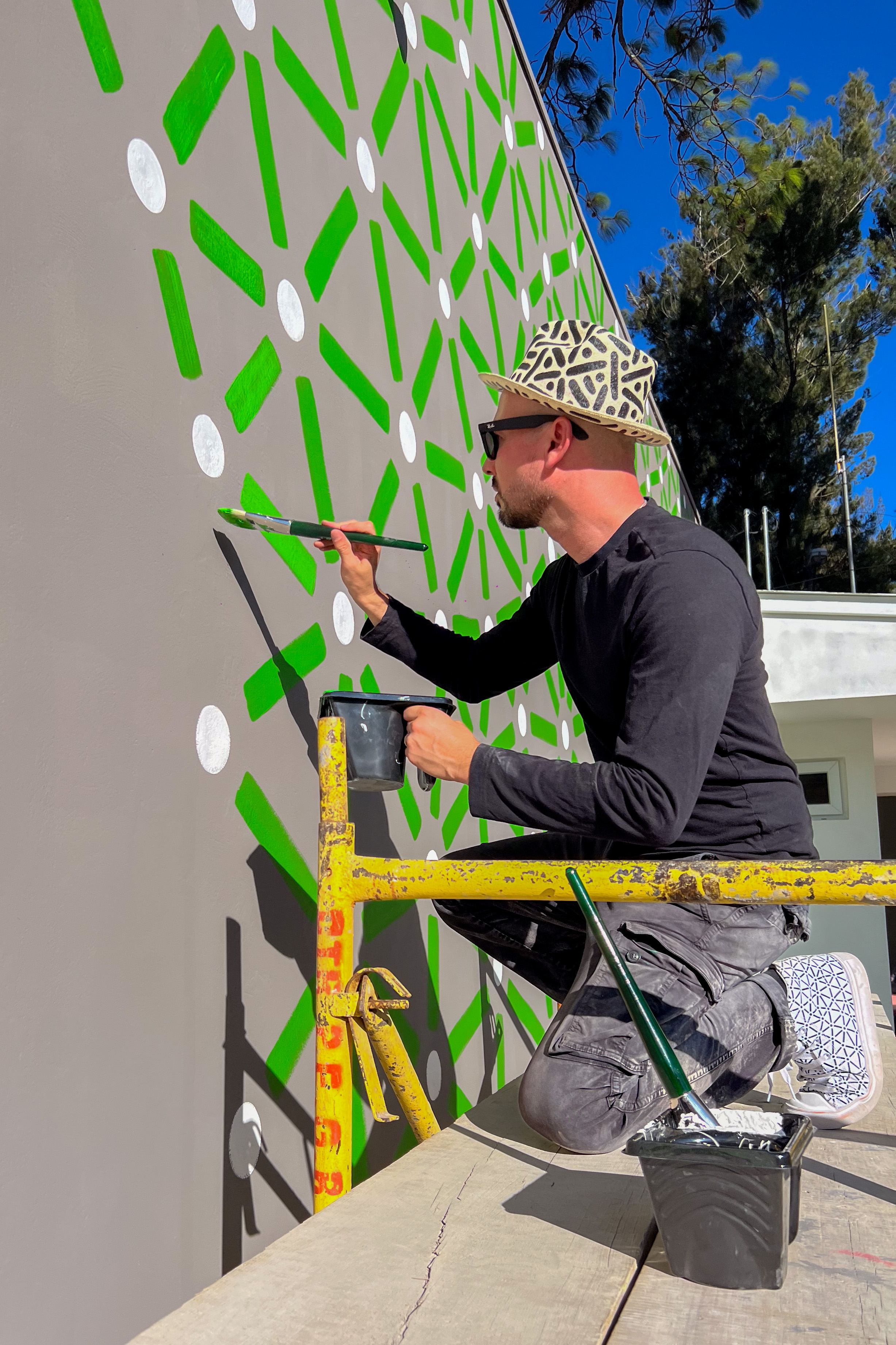 Roots: Artist sits on yellow scaffolding and paints green marks on a gray courtyard wall.