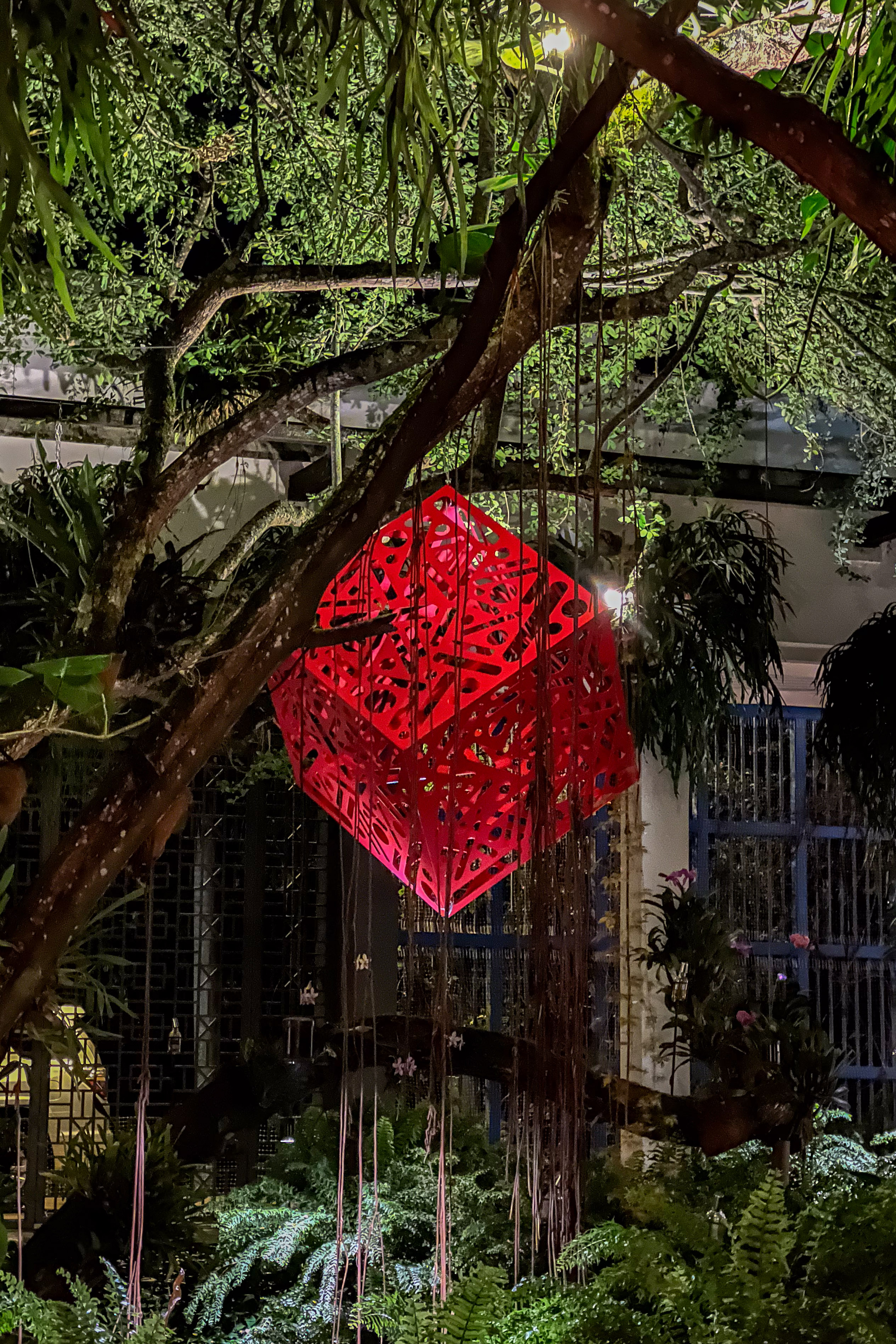 Hemoglobin: Red cube sculpture with patterned cutouts suspended among trees in a garden.