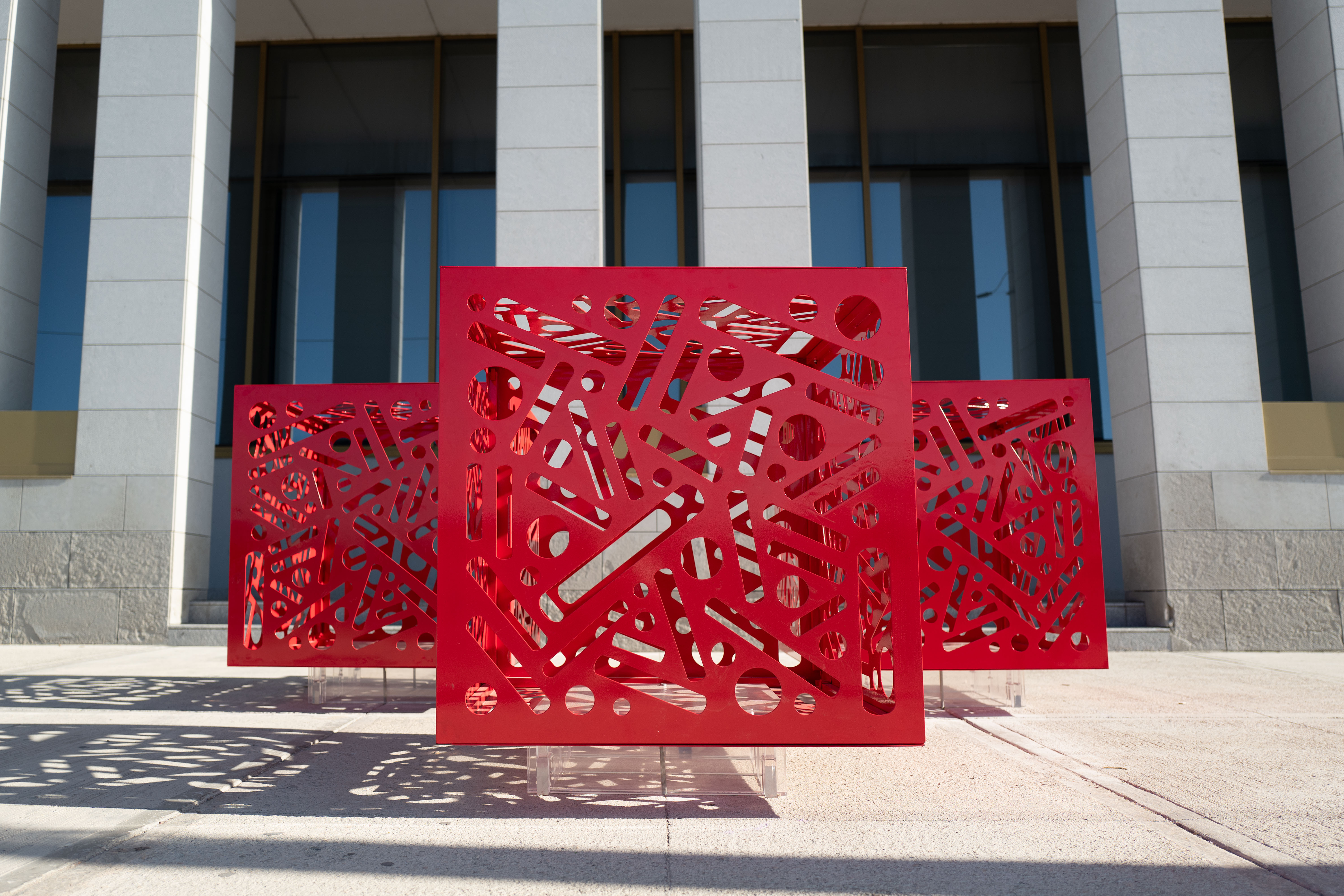 Hemoglobin - Installation of 3: Three red cutout cube sculptures cast geometric shadows onto sunlit pavement.