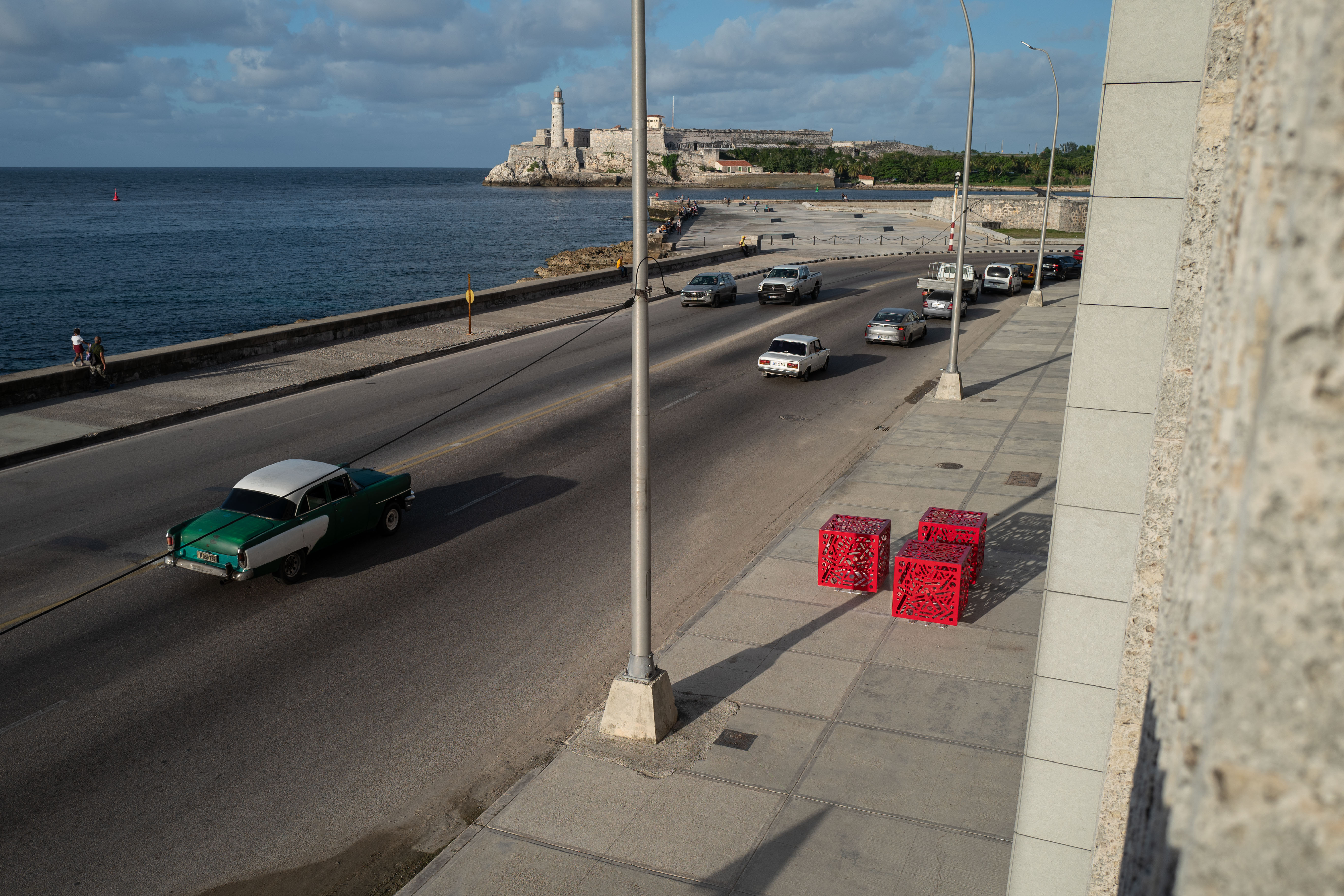 Hemoglobin - Installation of 3: Elevated view of three red cube sculptures beside a waterfront road.