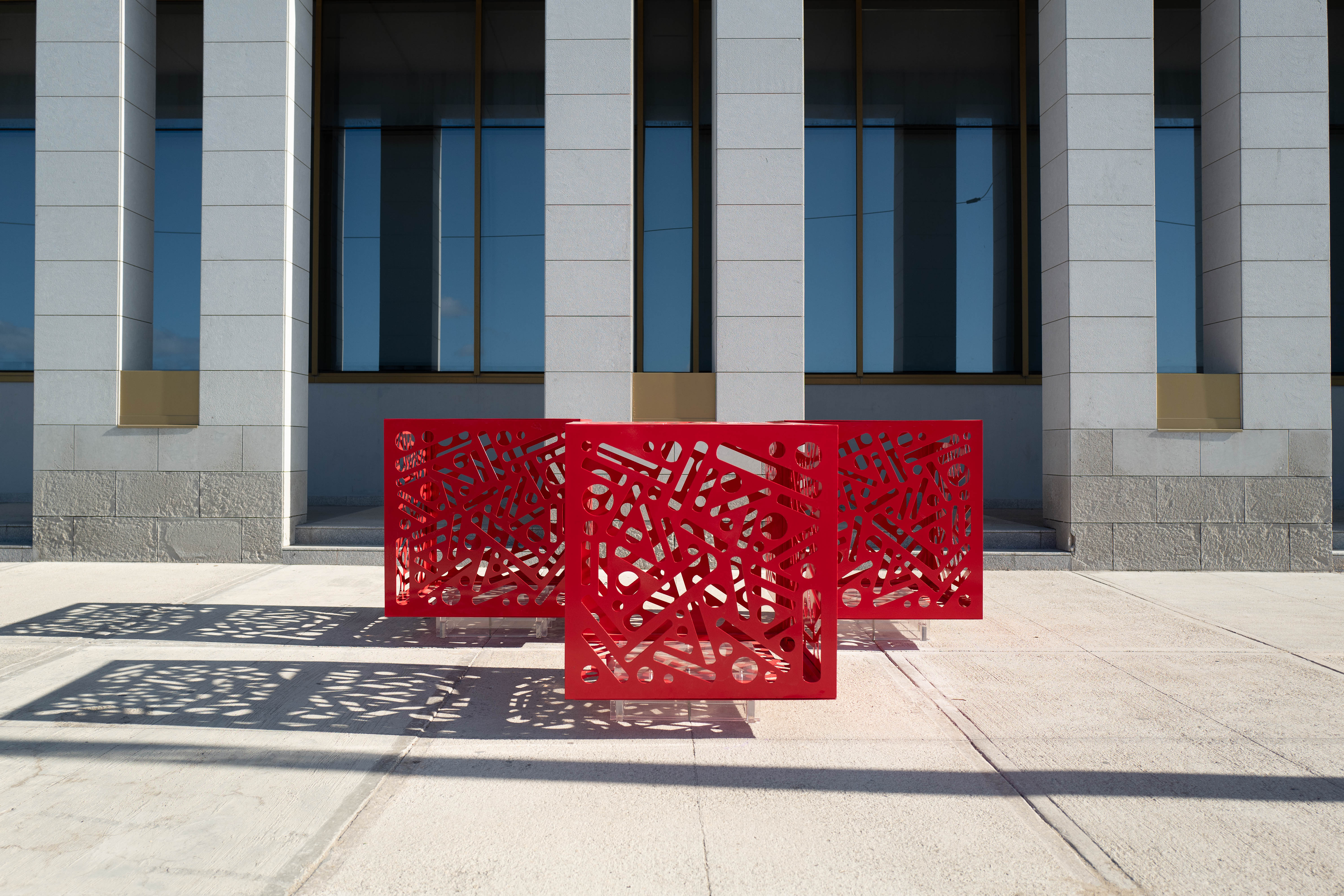 Hemoglobin - Installation of 3: Three red cutout cube sculptures cast geometric shadows onto sunlit pavement.