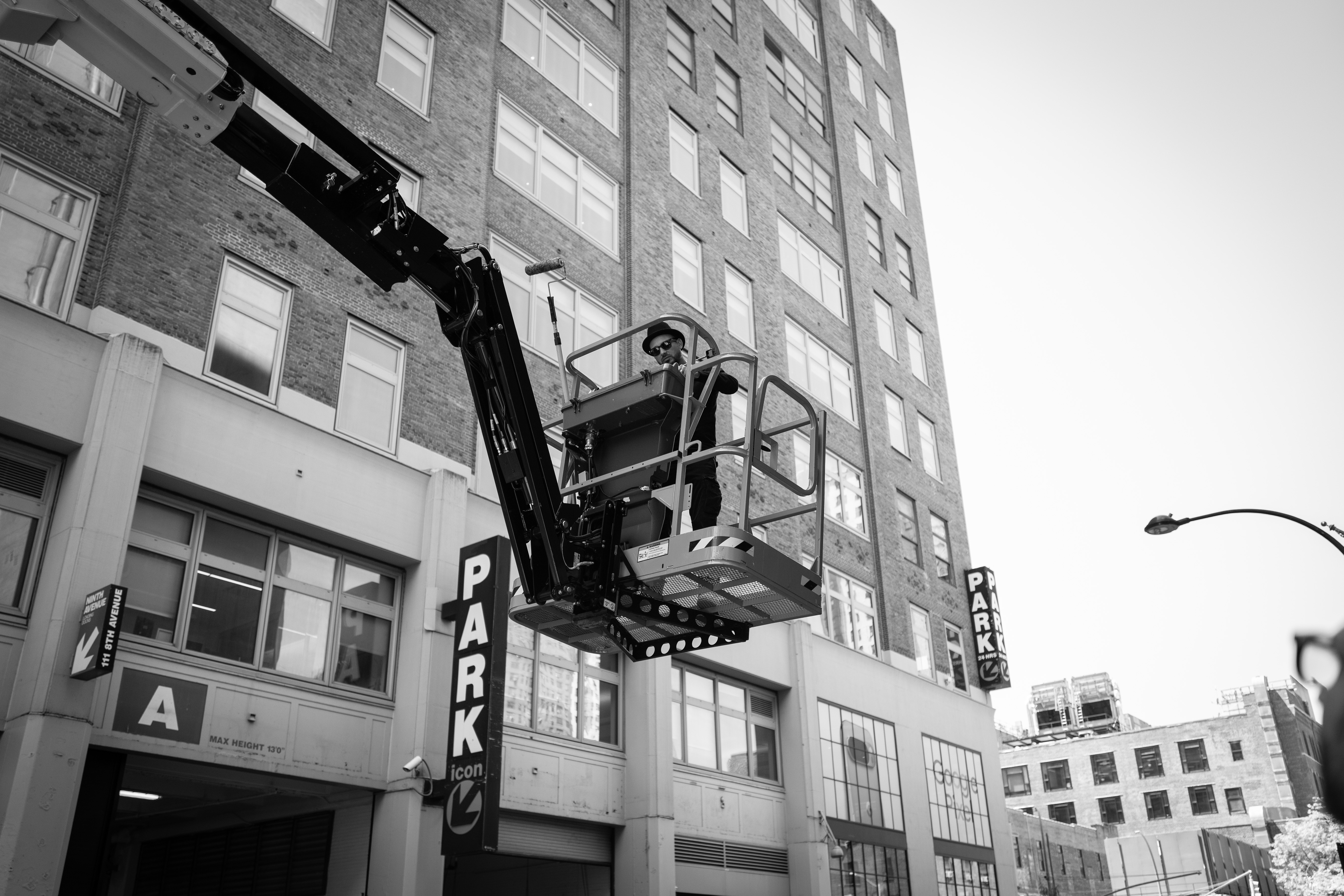 The Golden Weave: Black-and-white street view of an artist working from a raised lift beside a tall building.