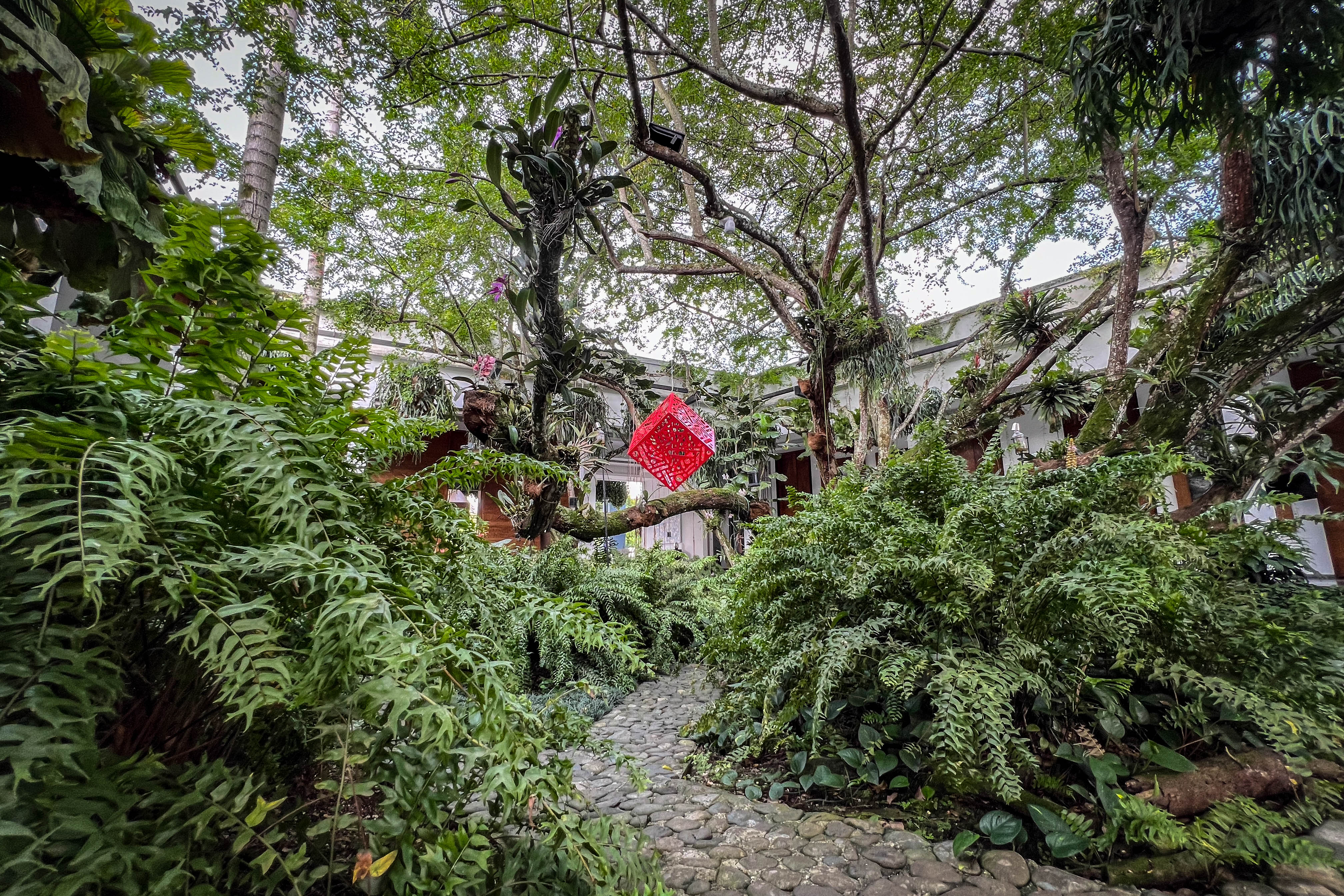 Hemoglobin: Red cube sculpture with patterned cutouts suspended among trees in a garden.